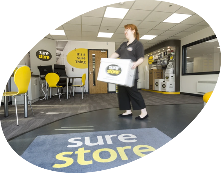 A staff member carrying a box in a modern office with yellow chairs and a carpet displaying the brand name 'sure store.' The office has a welcoming atmosphere with a clear view of branded signage.