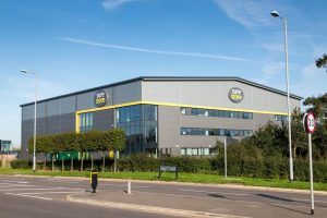 A modern storage facility named 'Sure Store' viewed from across the road, featuring a gray exterior with yellow accents and large windows, surrounded by landscaped greenery under a clear blue sky.