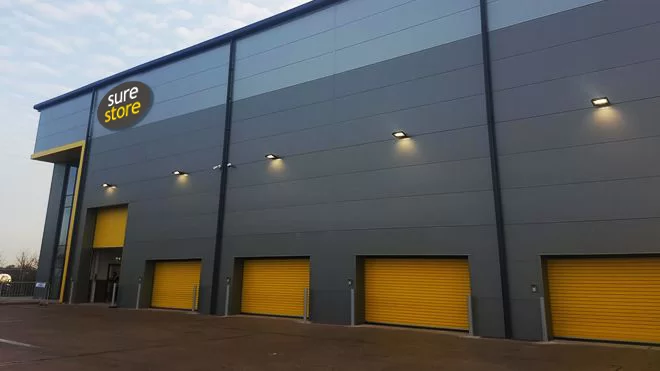 Exterior view of a modern storage facility named 'Sure Store.' The building features a gray facade with prominent yellow garage doors, under a cloudy sky.