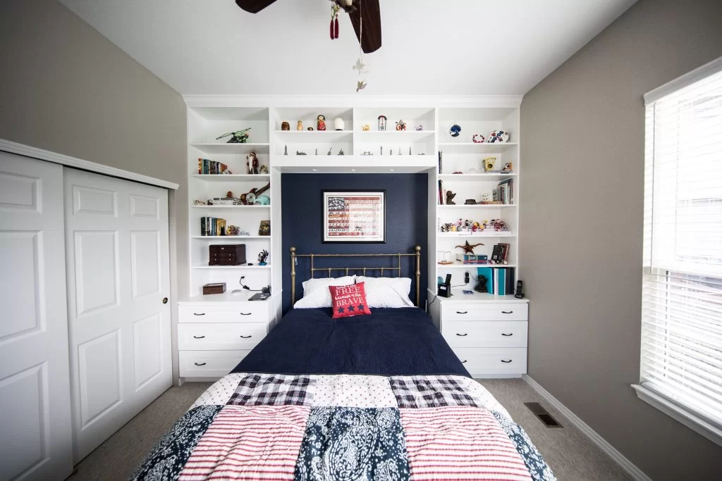 A neatly arranged bedroom featuring a dark blue bedspread on a bed with white pillows. Behind the bed, there is a blue wall with shelves displaying various items, and a framed American flag. White closet doors are visible on the left.