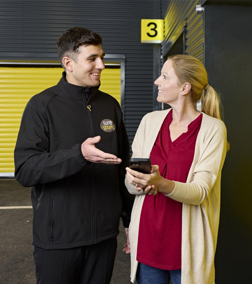 A man in a black jacket and a woman in a red top and cream cardigan are engaged in conversation. They stand beside a storage unit with yellow doors, with a number 3 visible on the wall.